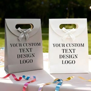 May include: Two gift bags, one gray and one white, with a handle and a bow. The bags have the text "YOUR CUSTOM TEXT DESIGN LOGO" printed on the front. Colorful ribbons are in the foreground.