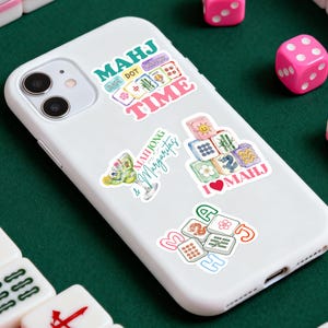 Może przedstawiać: Biały smartfon z wieloma kolorowymi naklejkami z motywami Mahjonga. Naklejki zawierają tekst, taki jak "MAHJ TIME" i "Mahjong & Margaritas". R&oacute;żowe kostki do gry są rozrzucone wok&oacute;ł telefonu na zielonej powierzchni.