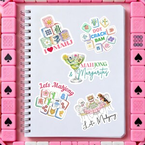 Może przedstawiać: Biały spiralny notatnik z kilkoma kolorowymi naklejkami przedstawiającymi kafelki Mahjong, koktajle oraz tekst "I ❤️ MAHJ", "Mahjong & Margaritas" i "Let's Mahjong". Naklejki otoczone są r&oacute;żowymi kafelkami Mahjong.
