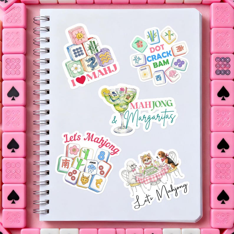 Może przedstawiać: Biały spiralny notatnik z kilkoma kolorowymi naklejkami przedstawiającymi kafelki Mahjong, koktajle oraz tekst "I ❤️ MAHJ", "Mahjong & Margaritas" i "Let's Mahjong". Naklejki otoczone są r&oacute;żowymi kafelkami Mahjong.