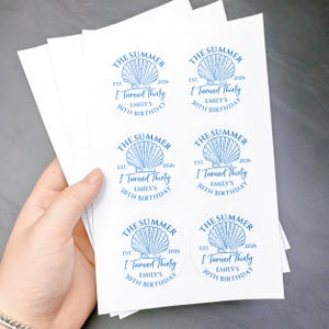 May include: A stack of white stickers with a blue seashell design and text. The text reads "THE SUMMER EST. 2026 I Turned Thirty EMILY'S 30TH BIRTHDAY". The stickers are round and are held in a person's hand.