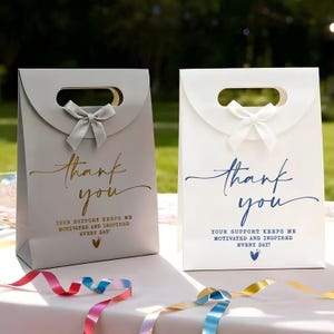 May include: Two gift bags, one gray and one white, with a handle and a bow. The bags have the words "thank you" in script, with the message "Your support keeps me motivated and inspired every day!"