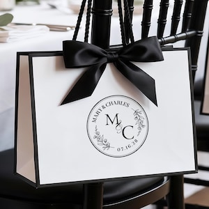 Puede incluir: Una bolsa de regalo blanca con bordes negros y asas de cuerda negra, con un gran lazo de satén negro en la parte superior. El centro muestra un gráfico circular negro con el texto MARY & CHARLES, las iniciales M & C y la fecha 07.16.28.