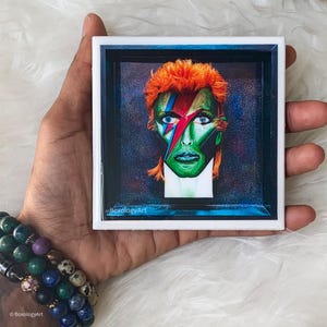May include: A white ceramic coaster with a blue border and a framed image of David Bowie with orange hair and colourful makeup. The image is set against a dark blue background with a glittery effect. The text "@BoxologyArt" is visible in the bottom left corner of the image.