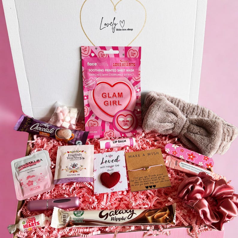 A white gift box filled with pink self-care items, including a sheet mask, lip balm, a brown fuzzy headband, a chocolate bar, and a small red heart.