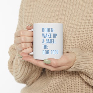 Könnte beinhalten: Eine weiße Keramiktasse mit blauem Text, der "OGDEN: WAKE UP & SMELL THE DOG FOOD" lautet.