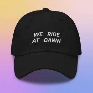 May include: Black baseball cap with the embroidered text "WE RIDE AT DAWN" in white. The cap has a curved brim and a rounded crown. The hat is made of a soft material and is suitable for casual wear.