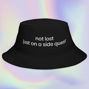 May include: Black bucket hat with white embroidered text that reads "not lost just on a side quest." The hat has a wide brim and a rounded crown. The background is a gradient of pastel colors.