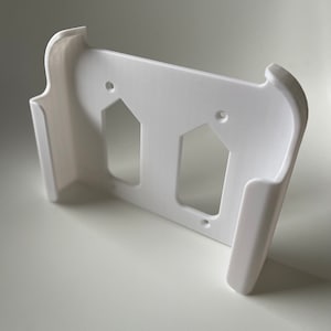 May include: White plastic wall mount with two cutouts for holding devices. The mount has rounded edges and a smooth finish.