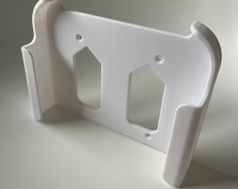 Puede incluir: Soporte de pared de plástico blanco con dos recortes para sujetar dispositivos. El soporte tiene bordes redondeados y un acabado liso.