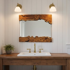 以下が含まれることがあります： ライブエッジの木製フレームの長方形の鏡が白い洗面台の上に吊り下げられています。 洗面台は、金色の蛇口とハンドルが付いた木製の洗面化粧台に設置されています。 白いガラスシェードの2つの壁取り付け用燭台が白い羽目板の壁に取り付けられています。