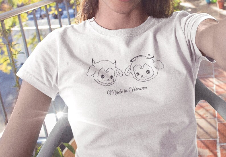 May include: White t-shirt with a graphic of two cartoon sheep, one with horns and the other with a halo. The text "Made in Heaven" is printed below the sheep.