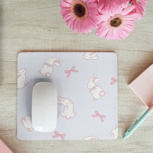 May include: A white computer mouse sits on a light purple mouse mat with a repeating pattern of white bunnies and pink bows. The mouse mat is on a light wood surface. A pink notepad and a light blue pen are on the right side of the image. A laptop computer and a bouquet of pink flowers are on the left side of the image.
