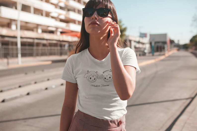 May include: White t-shirt with a black line drawing of two cartoon sheep, one with horns and one with a halo. The text "Made in Heaven" is below the sheep.