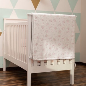 Op de afbeelding: Een wit babybedje met een roze bloemetjesdeken gedrapeerd over de bovenste leuning. Het babybedje staat in een kamer met een lichtgroen en gouden geometrisch wandpatroon.