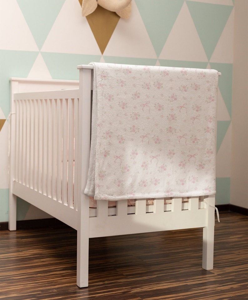 Op de afbeelding: Een wit babybedje met een roze bloemetjesdeken gedrapeerd over de bovenste leuning. Het babybedje staat in een kamer met een lichtgroen en gouden geometrisch wandpatroon.