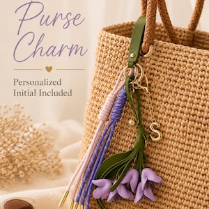 May include: A purse charm featuring a green leather strap, purple flower-shaped pendants, and long purple and light pink cord tassels with gold-tone metal tips. A gold-tone letter S charm hangs from a metal ring attached to a woven tan handbag.