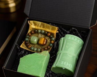 May include: A black gift box contains a green bar of soap, a bracelet with green and brown beads, and a bundle of green soap shaped like money. The box is lined with black crinkle paper.