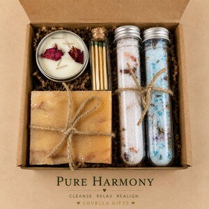 May include: A gift box containing a round candle with rose petals, a glass vial of matches, a square bar of soap tied with twine, and two glass test tubes filled with pink and blue bath salts. The text reads PURE HARMONY CLEANSE. RELAX. REALIGN. LOVELLA GIFTS.