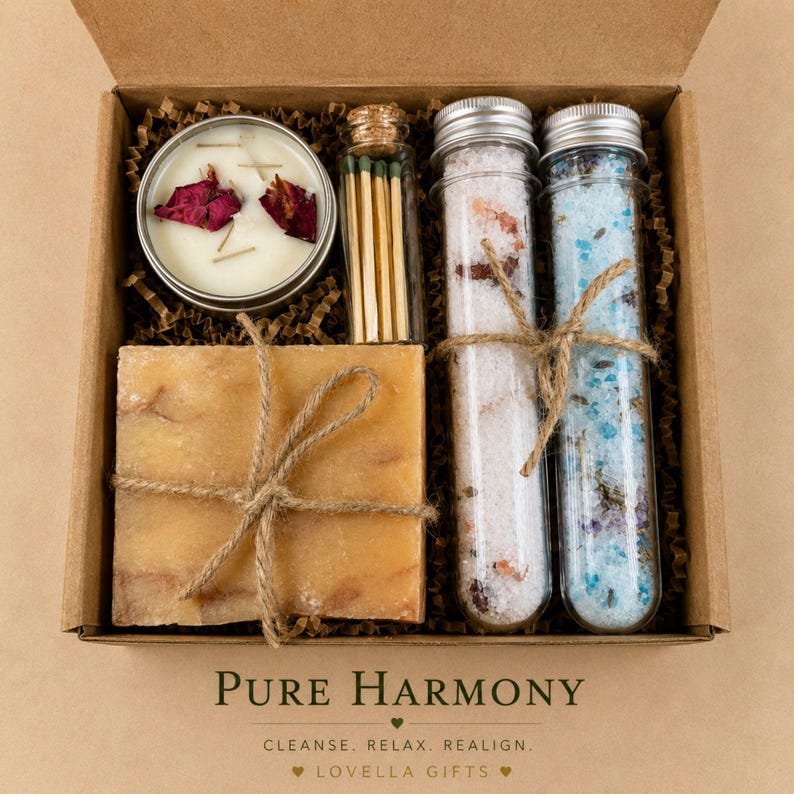 May include: A gift box containing a round candle with rose petals, a glass vial of matches, a square bar of soap tied with twine, and two glass test tubes filled with pink and blue bath salts. The text reads PURE HARMONY CLEANSE. RELAX. REALIGN. LOVELLA GIFTS.