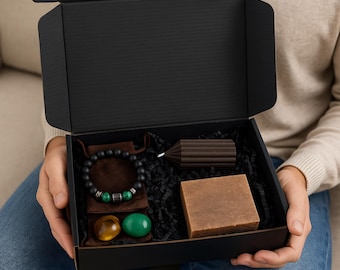May include: A black gift box containing a black beaded bracelet with green accents, two gemstones, a brown candle, and a brown bar of soap. The box is open, revealing the contents.