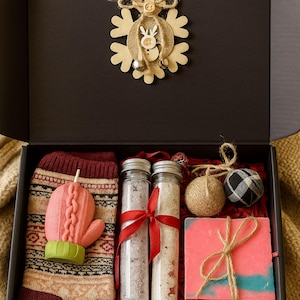 May include: A gift box containing a snowflake decoration, a patterned sock with a pink mitten, two glass tubes of bath salts, a pink soap bar, and twine balls. The box is black with the text "Lovella Gifts".