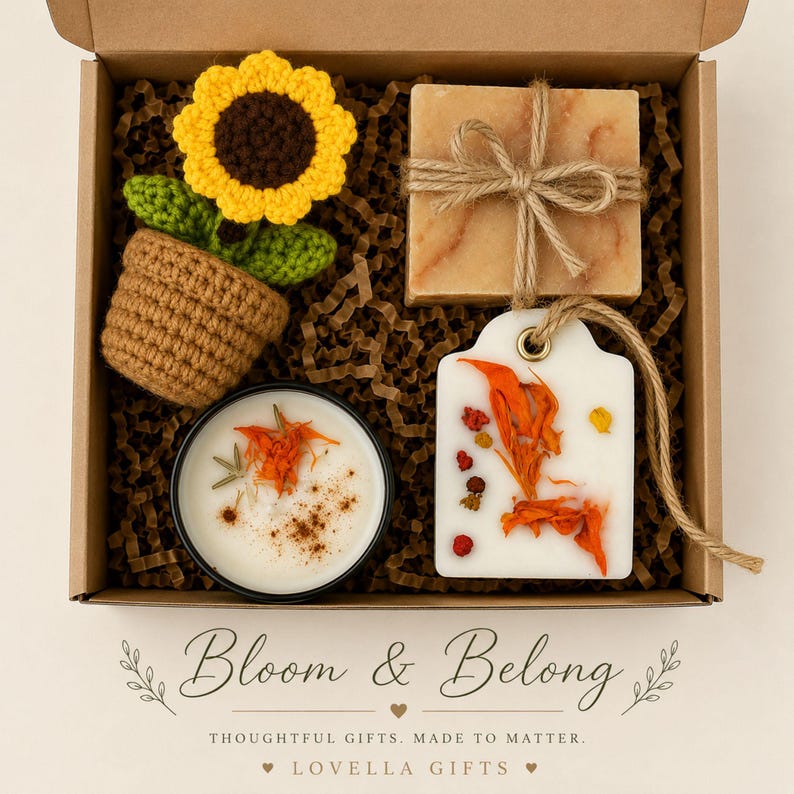 May include: A gift box containing a crocheted sunflower in a brown pot, a square bar of soap tied with twine, a round candle with botanical toppings, and a white wax melt tag decorated with dried orange petals and small red floral accents.