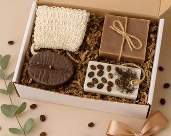 May include: A gift box with coffee-themed items. It contains a beige washcloth, a coffee bean-shaped soap, a brown soap bar tied with twine, and a white soap bar decorated with coffee beans. Scattered coffee beans and a ribbon are also present.