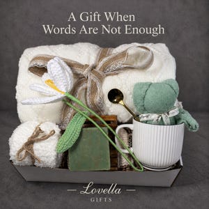 Peut inclure: Un panier cadeau avec une couverture blanche nouée avec un ruban de jute, une tasse blanche avec un ours en peluche vert, une fleur au crochet, un pain de savon et une cuillère dorée. Le texte "A Gift When Words Are Not Enough" est en haut.