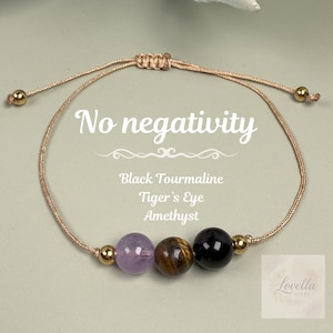 May include: Adjustable bracelet featuring three gemstone beads: amethyst, tiger's eye, and black tourmaline. The bracelet is strung on a beige cord with gold-colored accents. Text on the image reads "No negativity".