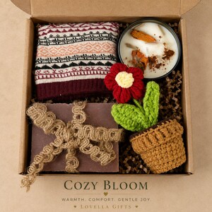 May include: A gift box containing a folded maroon and cream patterned knit fabric, a round candle in a black tin with dried orange slices, and a crocheted red flower in a brown pot. A dark rectangular item is wrapped in decorative jute ribbon.