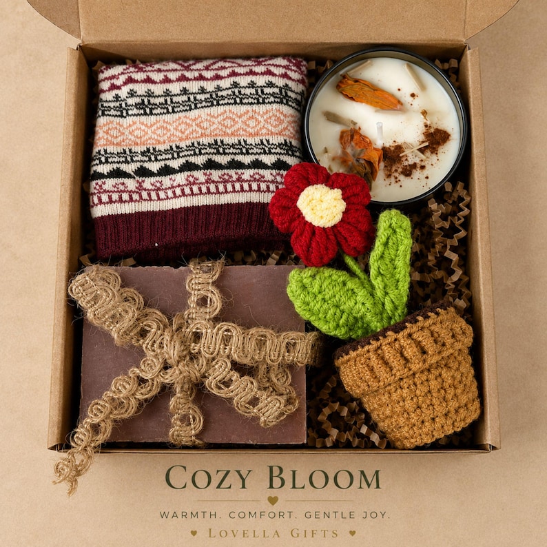 May include: A gift box containing a folded maroon and cream patterned knit fabric, a round candle in a black tin with dried orange slices, and a crocheted red flower in a brown pot. A dark rectangular item is wrapped in decorative jute ribbon.