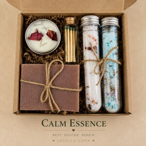 May include: A gift box containing a round candle with rose petals, a glass vial of matches, two glass test tubes filled with bath salts, and a rectangular brown soap bar tied with twine. The box features the text CALM ESSENCE REST. SOOTHE. RENEW.