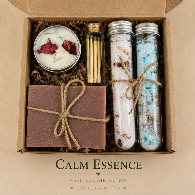 May include: A gift box containing a round candle with rose petals, a glass vial of matches, two glass test tubes filled with bath salts, and a rectangular brown soap bar tied with twine. The box features the text CALM ESSENCE REST. SOOTHE. RENEW.