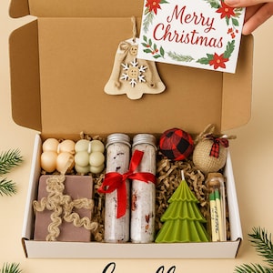 May include: A Christmas-themed gift box filled with festive items. Includes a bell ornament, a card with "Merry Christmas" text, candles, bath salts, a tree-shaped candle, and other decorative elements. The box is labeled "Lovella Gifts".