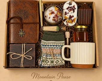 May include: A gift box containing a brown leather journal with a compass design, a gold pen, a tin of bath salts, a floral wax melt, a brown candle, a glass vial of matches, a bar of soap, patterned socks, honey, a dipper, and a ribbed mug.