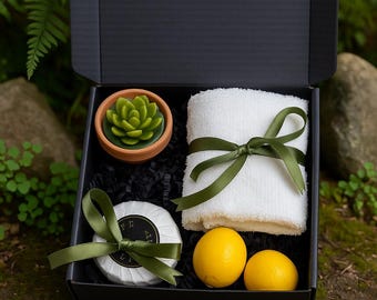 May include: A black gift box containing a green succulent-shaped candle in a terracotta pot, a white towel tied with a green ribbon, two yellow lemons, and a round soap with a green ribbon.