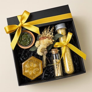 Op de afbeelding: Een zwarte geschenkdoos met een gele strik bevat een groene kaars, gedroogde bloemen, een gele zeep en glazen flesjes gevuld met gele korrels. De doos is gevoerd met zwart snipperpapier.