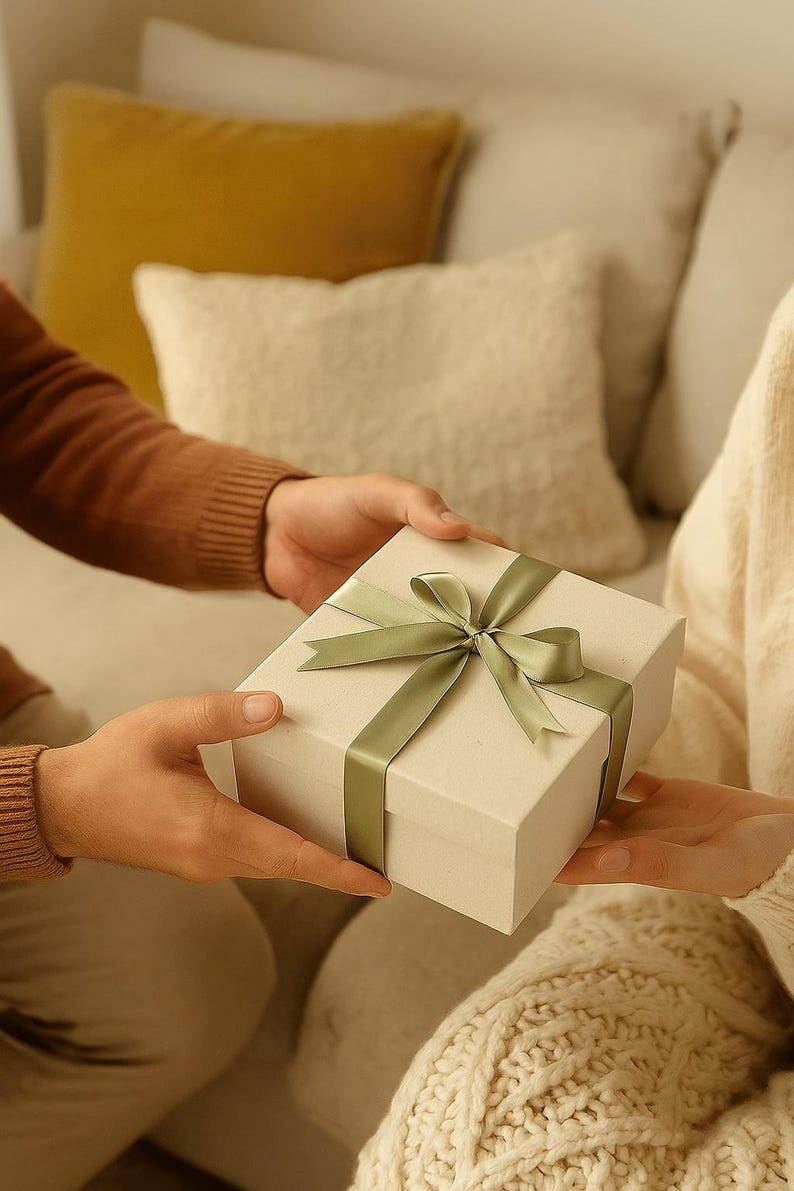 Op de afbeelding: Een rechthoekige, cr&egrave;mekleurige geschenkdoos met een saliegroen lint wordt overhandigd. De achtergrond toont een bank en kussens. De sc&egrave;ne toont een persoon die een cadeau aan een ander geeft.