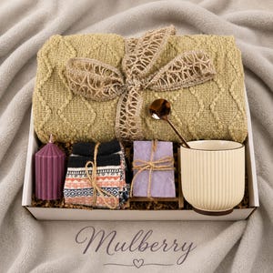 May include: A gift box containing a folded olive green cable-knit blanket tied with a burlap ribbon, a purple ribbed pillar candle, patterned socks, a bar of purple soap on a wooden rack, a gold spoon, and a cream-colored ribbed ceramic mug.