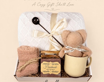 May include: A self-care gift box with a white blanket, a teddy bear in a beige mug, a bar of soap, and a pair of pink socks. The box is tied with a cream ribbon and includes a black spoon. Text on the box reads "Self Care Gift Box".