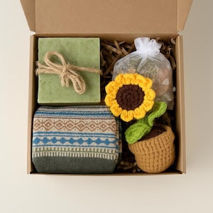 May include: A gift box filled with a green bar of soap tied with twine, a pair of patterned socks, a crocheted sunflower, and a small bag of potpourri. The box is made of cardboard and filled with brown paper.