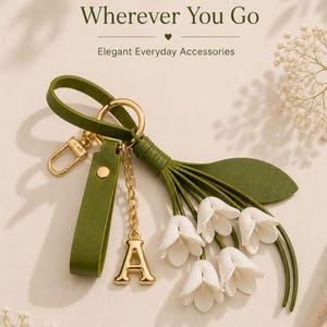 May include: A green leather keychain with a gold-colored clasp and chain, featuring a gold letter "A" charm and a cluster of white flower accents. The text "Bloom Wherever You Go" is at the top.