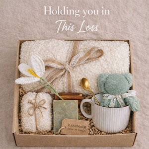 May include: A gift box with a cream-colored towel tied with a ribbon, a white crochet flower, a small green teddy bear in a white mug, soap, and a gold spoon. The box includes the text "Holding you in This Loss."