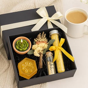 Op de afbeelding: Zwarte geschenkdoos met een cr&egrave;mekleurig lint, met een groene roosvormige kaars, een gele zeep, gedroogde bloemen en glazen flesjes met gele inhoud. Een witte mok met koffie en een kleine vaas met bloemen staan in de buurt.