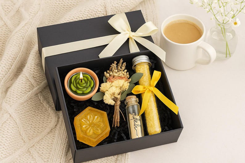 Op de afbeelding: Zwarte geschenkdoos met een cr&egrave;mekleurig lint, met een groene roosvormige kaars, een gele zeep, gedroogde bloemen en glazen flesjes met gele inhoud. Een witte mok met koffie en een kleine vaas met bloemen staan in de buurt.