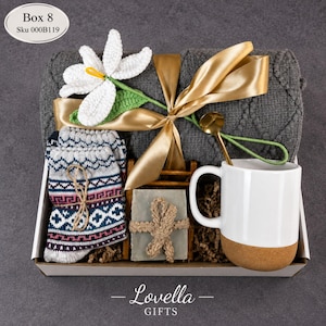 May include: A gift box with a gray blanket, white mug with cork base, patterned socks, soap, and a crochet flower. The box is tied with a gold ribbon. The text "Lovella GIFTS" is at the bottom.