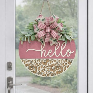Puede incluir: Un letrero de puerta redondo de madera rosa con la palabra "hello" en cursiva. El letrero presenta una sección inferior con estampado floral y está rematado con un lazo y vegetación. El lazo tiene estampados de cuadros rosas y blancos y florales.
