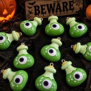 May include: A collection of green potion bottle ornaments with a single eyeball design. The Halloween-themed decorations are accented with white dots and a purple iris. A lit candle, pumpkins, and a sign that says "BEWARE" are in the background.