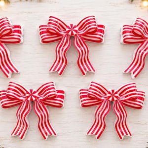 May include: Five decorative bows featuring a red and white candy cane striped pattern. Each bow has a central circular accent resembling a peppermint candy. The bows are arranged in a group, displaying a wavy ribbon texture and a glossy finish.